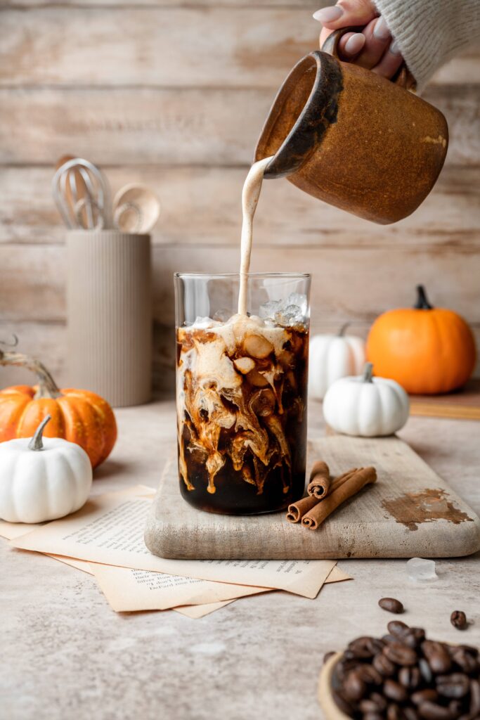 Cold brew coffee with a pumpkin spice creamer being poured into the glass. There are white and orange pumpkins scattered throughout the scene.