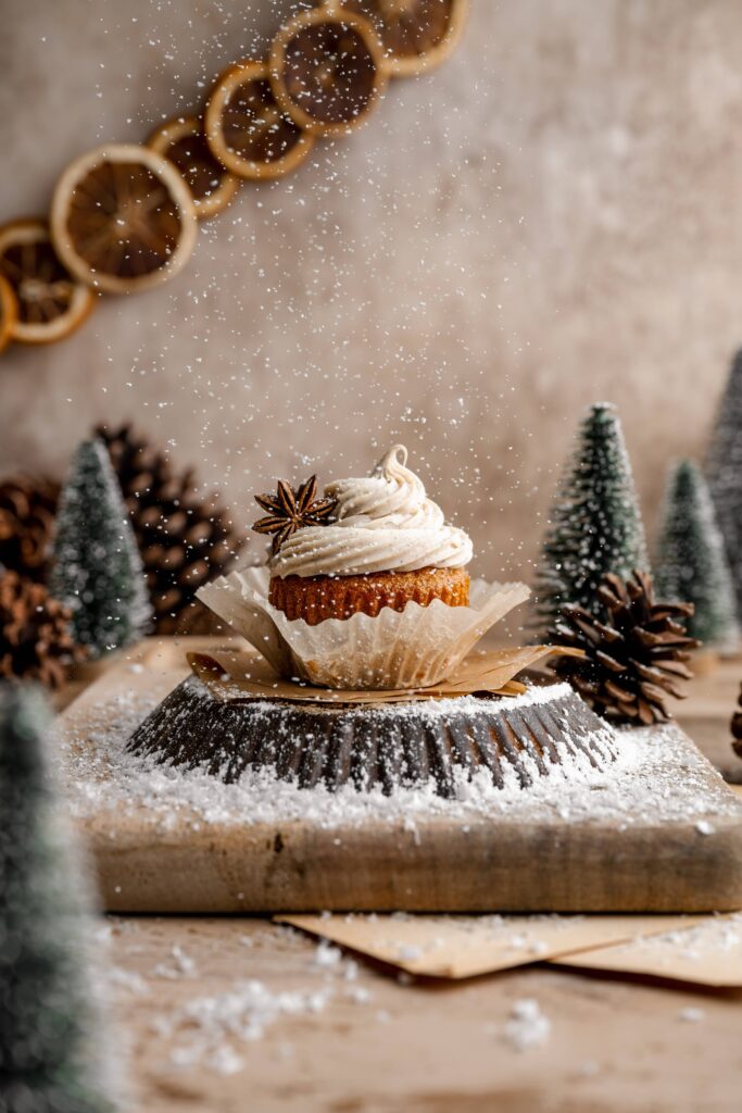 A spice cupcake with cream cheese frosting & a star anise garnish. There's powdered sugar snow throughout the scene.