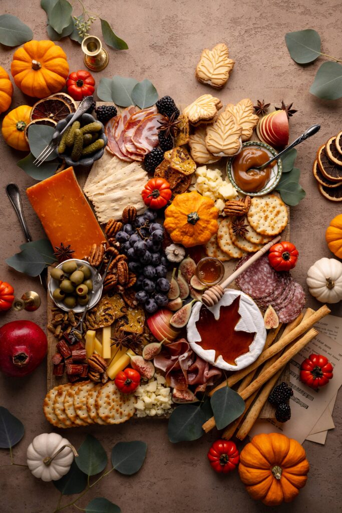 A Thanksgiving-inspired charcuterie board with pumpkin, maple leaf cream cookies, brie cheese, crackers, & salami.